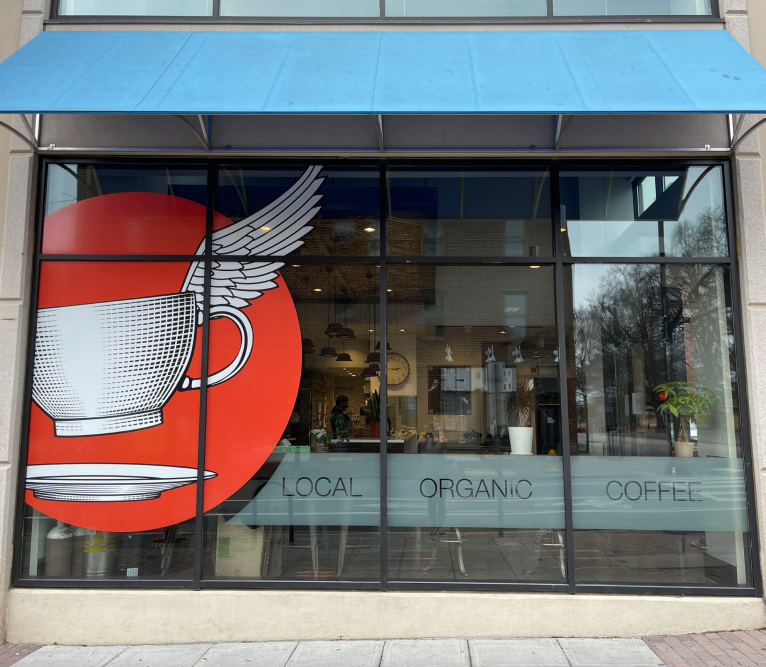 branding design in large storefront windows of a downtown raleigh coffee shop with bold graphics including a huge orange circle with a coffee cup with wings floating off of a saucer. There is also a bar with frosted vinyl that says "local, organic, coffee".